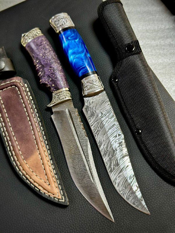 Два ножа с фиолетовой и синей рукоятками, видны дамасковые рисунки на лезвиях и коричневые кожаные ножны рядом.