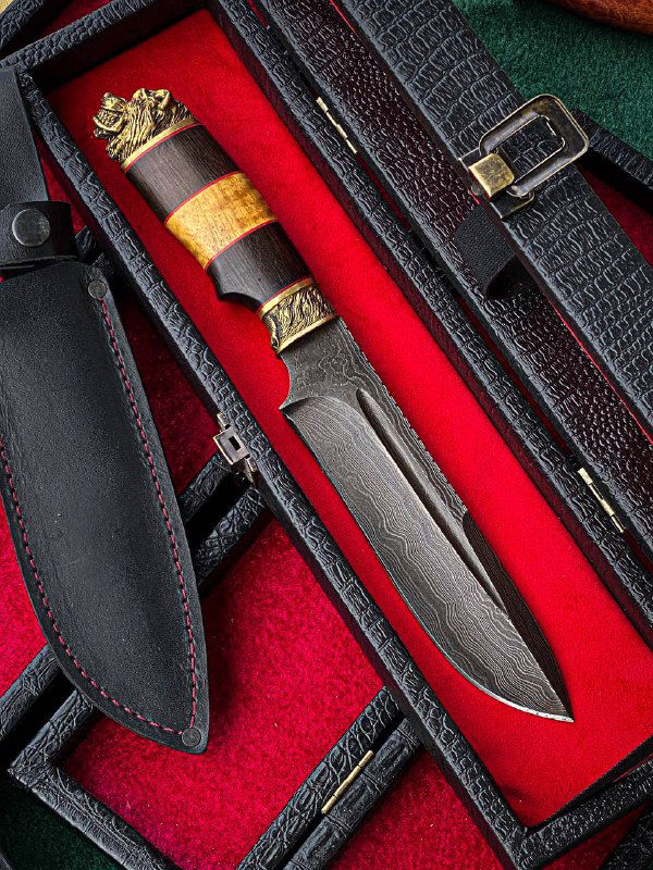 Нож с рукоятью из тёплой карельской берёзы и декоративными элементами на гардe, уложенный в футляр с видимыми ножнами.