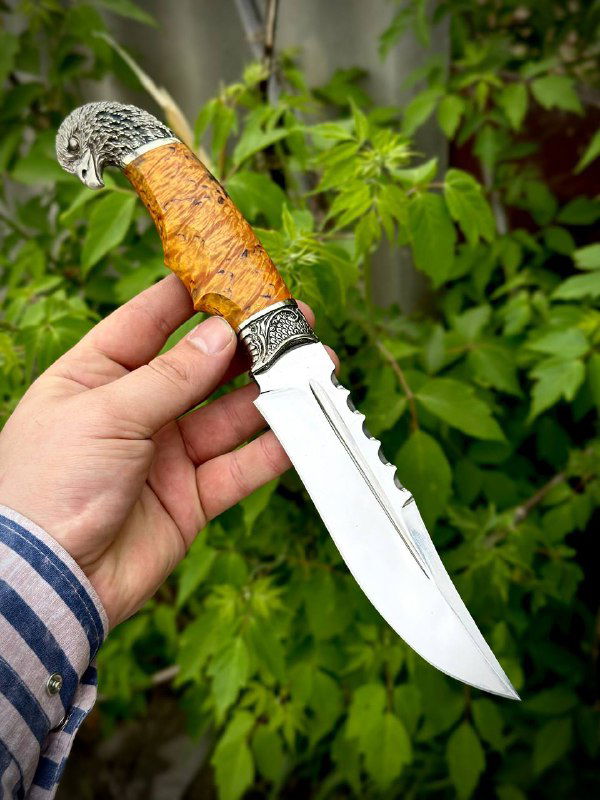 Нож модели «Орлан» в руке мастера: клинок из стали 95х18 и рукоять из стабилизированной карельской берёзы жёлтого оттенка на фоне листвы.