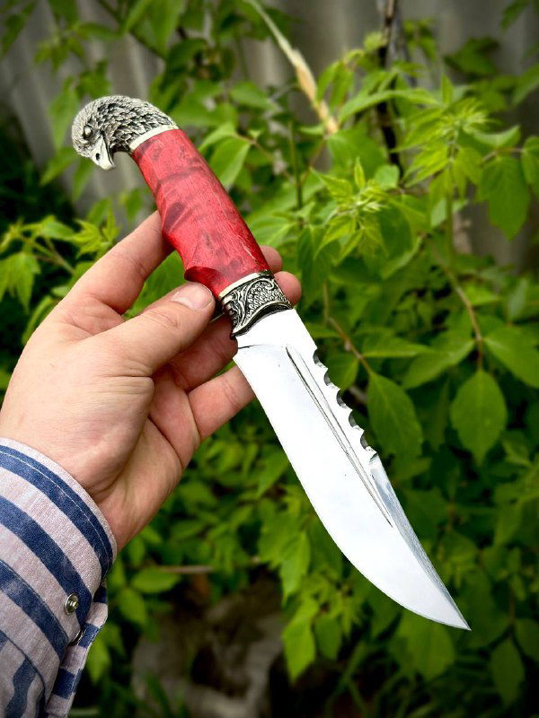 Нож модели «Орлан» с красной стабилизированной карельской рукоятью и характерным профилем клинка на фоне зелёной растительности.
