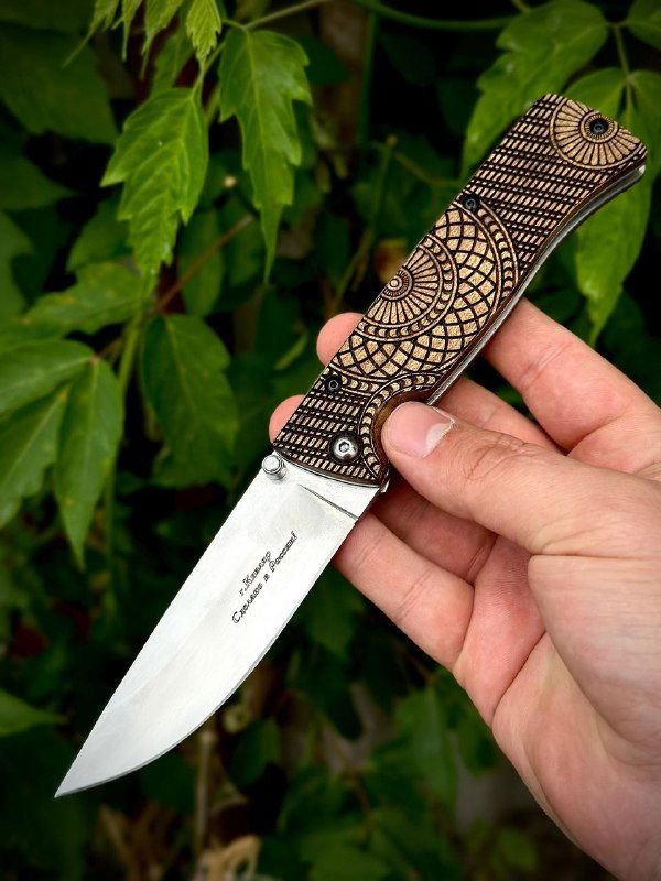 Фотография ножа «Шмель» в раскрытом состоянии: стальной клинок с гравировкой, декоративная рукоять из ореха, фон — листва на природе.