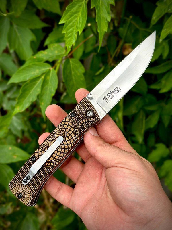 Складной нож «Шмель» в руке под углом: видна длина клинка, замок и декоративная картина рукояти на фоне зелёных листьев.