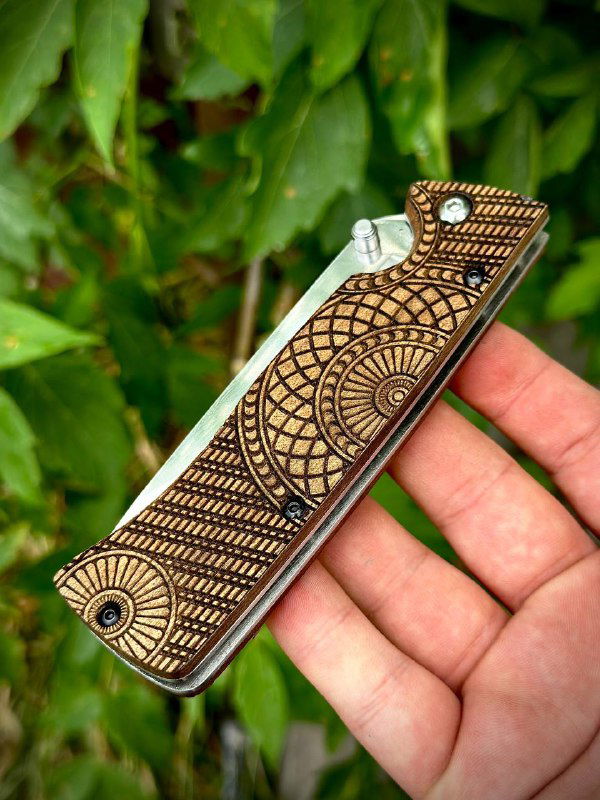 Фото сложенного ножа «Шмель»: ореховая рукоять с подробной художественной вырезкой и металлизированные элементы, снято крупно на зелёном фоне.