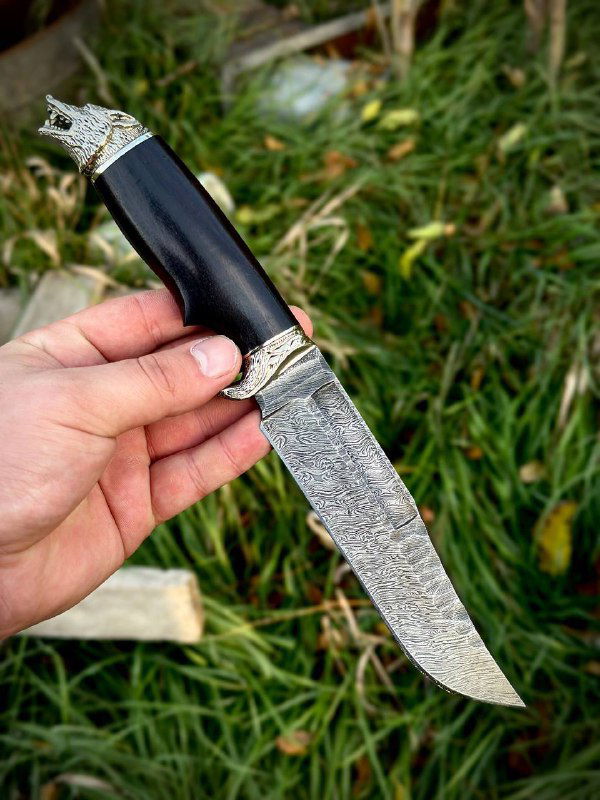 Крупный кадр рукояти ножа «Волк»: полированная тёмная рукоять из граба с декоративным навершием в виде головы волка из мельхиора на травяном фоне.