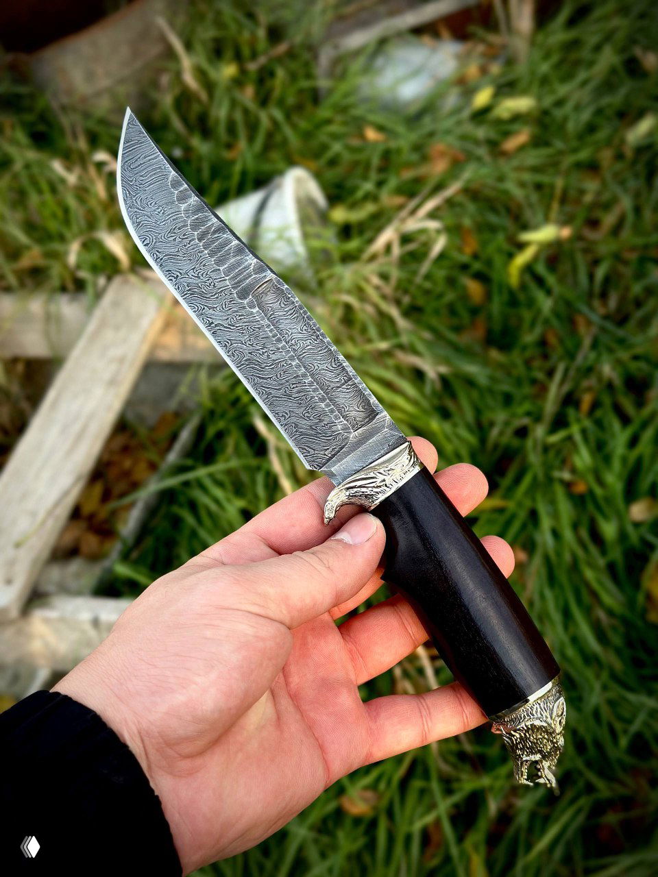 Клинок ножа «Волк» крупным планом: выразительный рисунок дамаска по всей длине лезвия, видна текстура и художественная обработка металла на фоне травы.