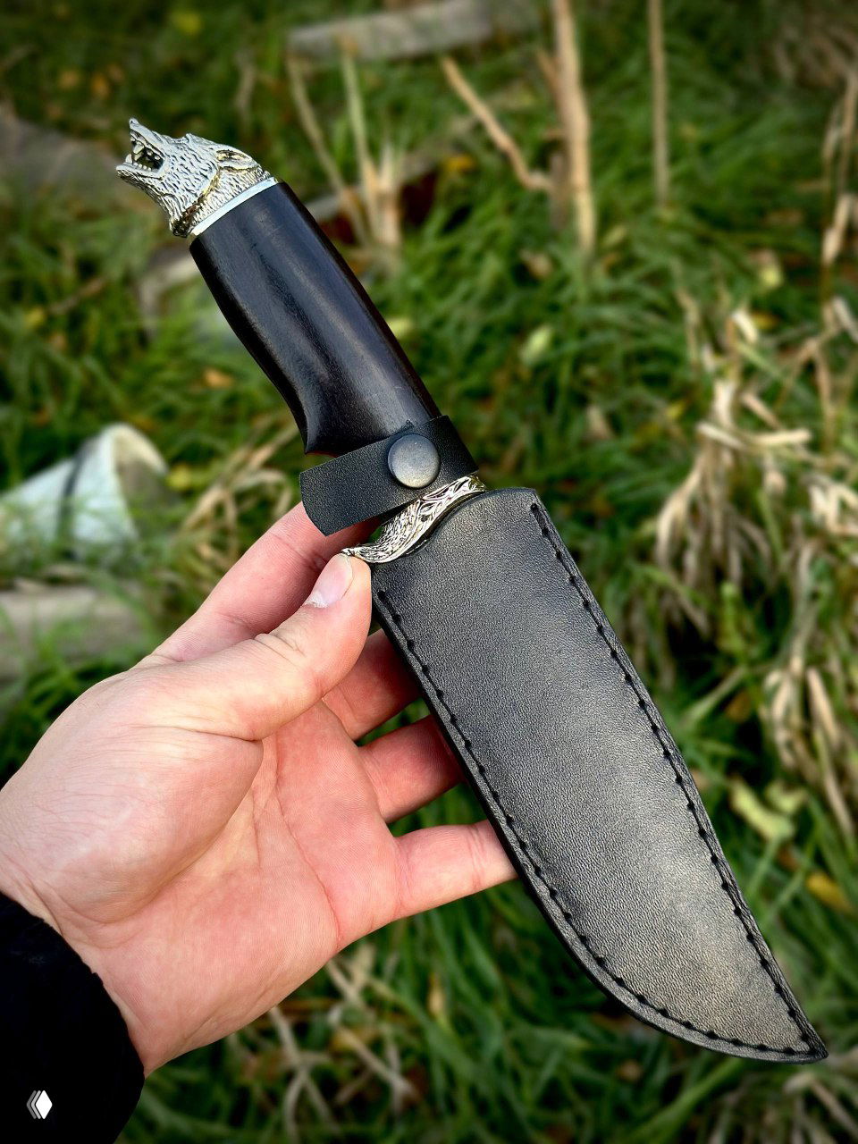 Ножны из кожи вместе с ножом «Волк»: тёмные кожаные ножны с кнопкой фиксации, гарда и часть рукояти видны на фоне природной травы.