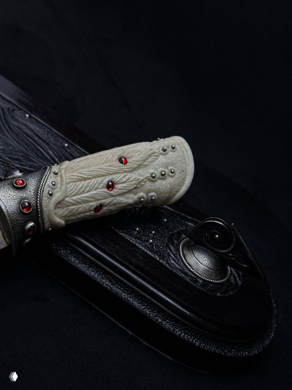 Деталь рукояти из бивня моржа с резьбой в виде пера и красными фианитами на фоне чёрной подставки; вид с близкого расстояния.