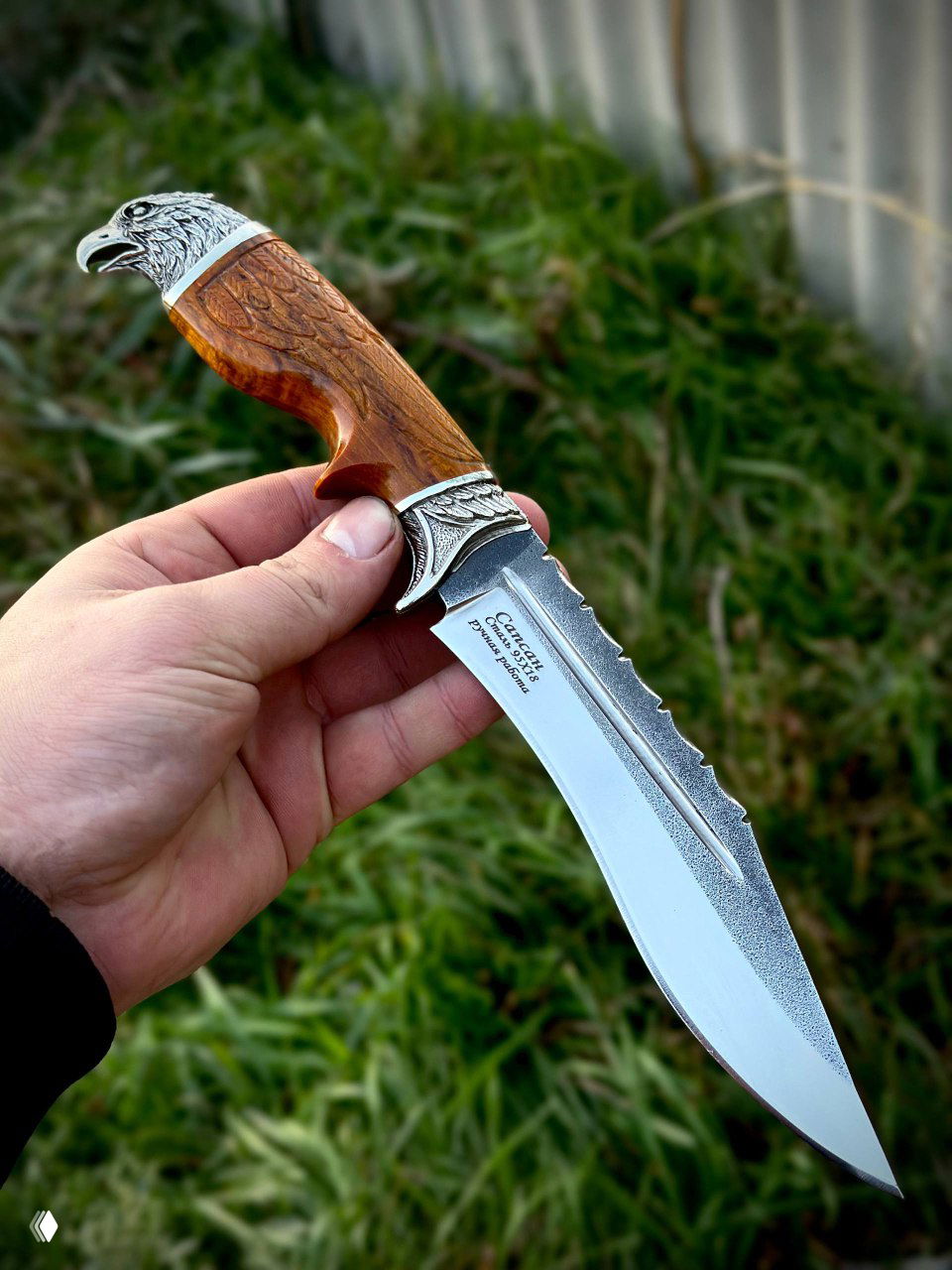 Близкий план ножа «Сапсан» с коричневой рукоятью из карельской берёзы, резными деталями и отполированным клинком на фоне травы; изделие держат в руке.