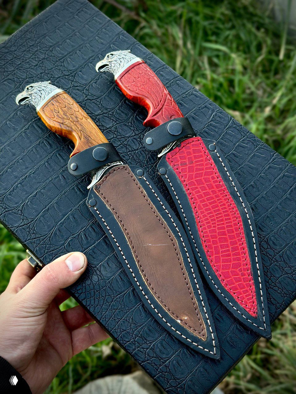 Два ножа в кожаных ножнах — красной и коричневой — уложены на черном фактурном чехле; видны декоративные элементы, швы и ручки ножен.