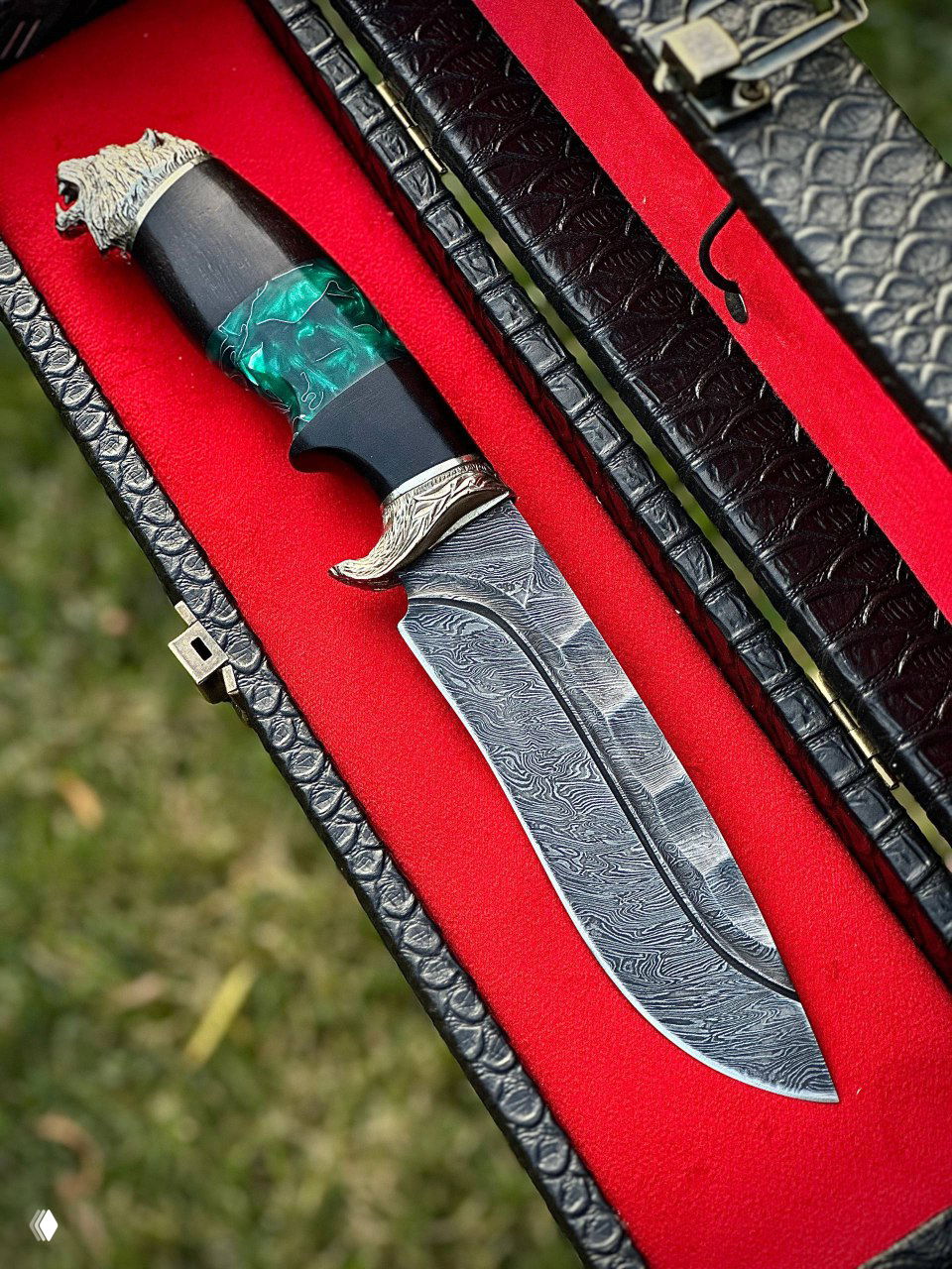 Крупный план ножа с дамасским клинком и зелёной акриловой вставкой в рукояти, размещённого в футляре с красной тканевой подкладкой, вид сверху.