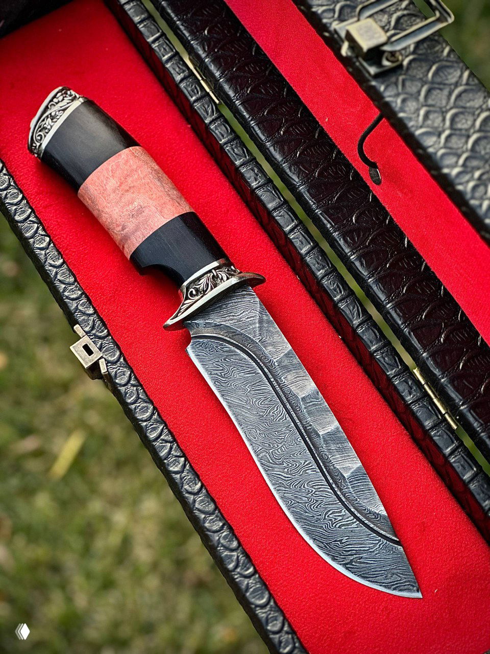 Крупный план второго ножа с дамасской текстурой клинка и рукоятью из карельской берёзы, лежащего в чёрном кейсе с красной подкладкой на зелёном фоне.