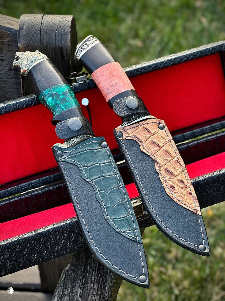 Две кожаные ножны в комплекте — тёмно-зелёная и светло-коричневая вставка под кожу крокодила, закреплённые на чёрном фоне кейса, рядом видны части рукоятей ножей.