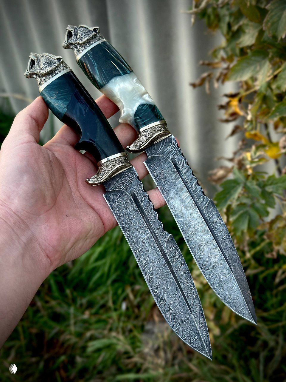 Два дамасских ножа в руках мастера: крупный план клинков с выраженным рисунком дамаска и мельхиоровыми гардами на фоне зелени