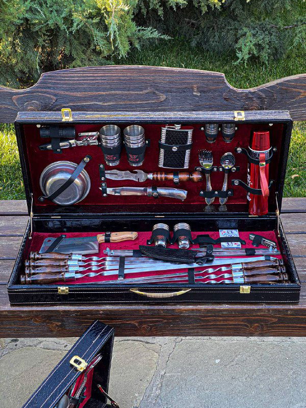 Общий вид набора в закрытом на защёлки кейсе, размещённый на столе на фоне зелёных кустов; заметны термос, фляжка и набор столовых приборов.