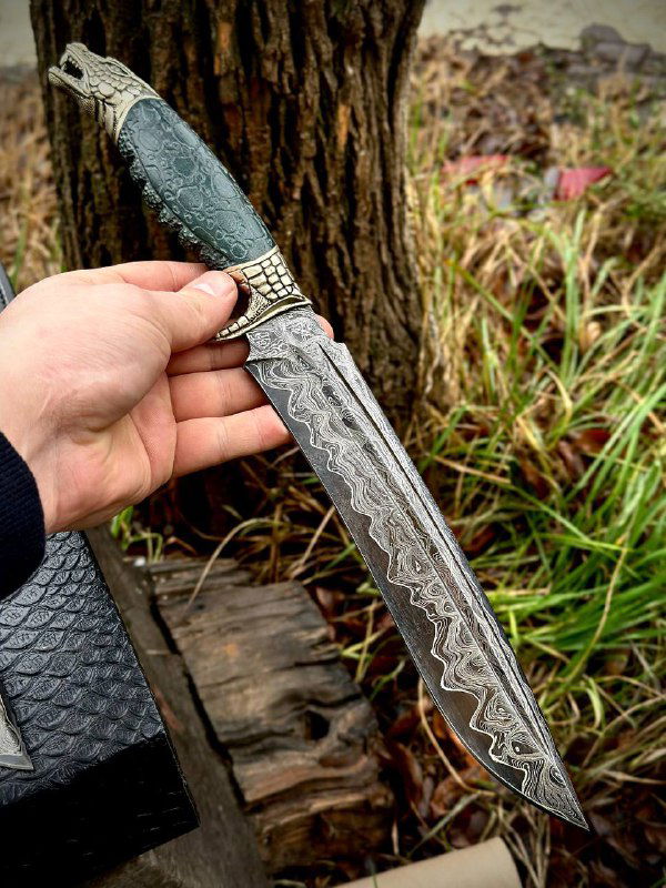 Кинжал «Дракон» в руке: видно лезвие с рисунком дамаска, мельхиоровая гарда и тёмно-зелёная рукоять из карельской берёзы на природном фоне.