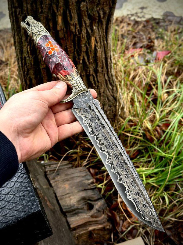 Кинжал «Дракон» в руке с красно-чёрной рукоятью в узор «соты» из акрила; отчётливо виден рисунок дамаска и мельхиоровая гарда.