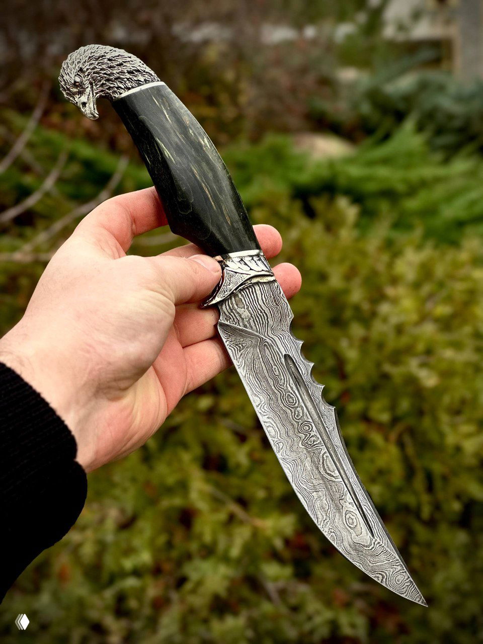 Крупный план ножа «Орлан» в руке: видна текстура дамаска на лезвии и тёмно-зелёная карельская рукоять с орлиной гардой.