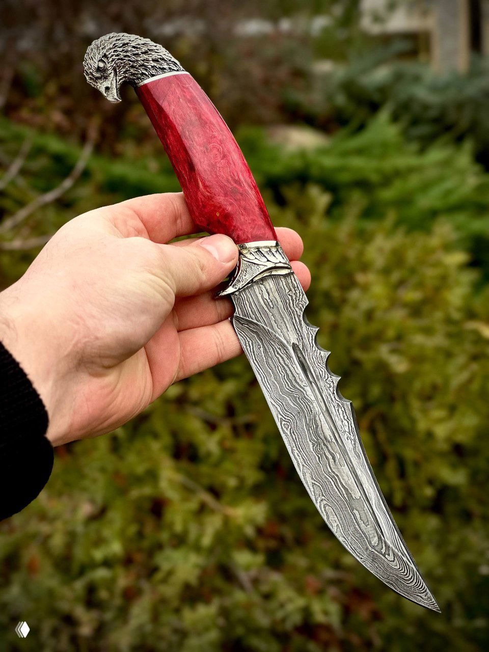 Красная рукоять ножа «Орлан» в ладони мастера: видна волнистая структура дамасской стали и фигурная гребёнка на обухе клинка.