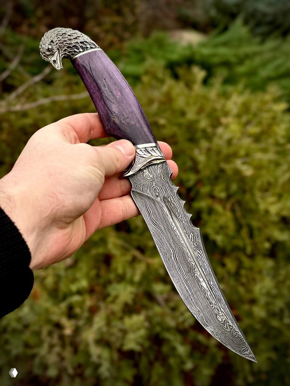 Фиолетовая рукоять ножа «Орлан» в руках: крупный план клинка с типичным дамасским рисунком и декоративной гардой в виде головы орлана.