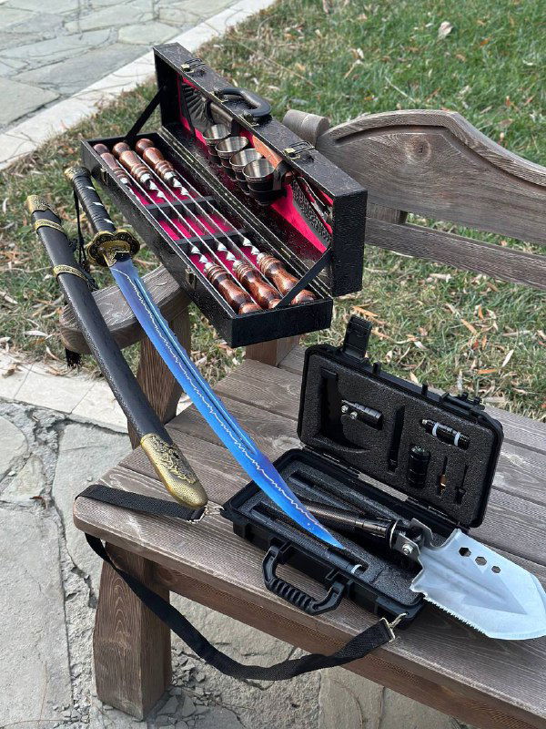 На скамье раскрыт кейс с шампурами и инструментами; рядом меч с синей ножной, складная лопата и многофункциональный подарочный набор