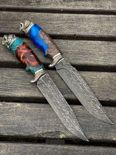 Два дамасковых ножа рядом на деревянной поверхности; крупный план лезвий с рисунком стали и разноцветных рукоятей, без людей на фото.