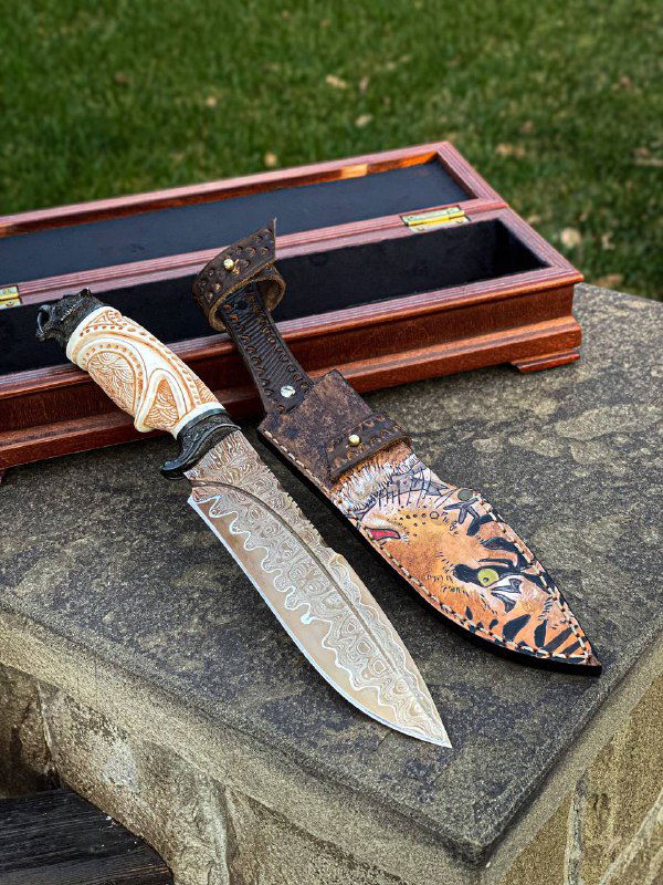 Нож и ножны на каменной поверхности с открытым деревянным кейсом: видно сочетание белой рукояти, мельхиоровых гард и расписного чехла.
