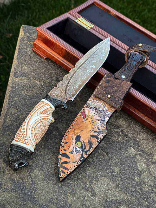 Вид под углом на авторский клинок: видно оксидирование, дамасскую структуру, чёрные гарды и рукоять с орнаментом на фоне зелёного газона.