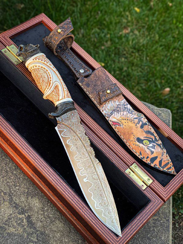 Фотография раскрытого набора: нож с дамасским клинком, резная рукоять из рога лося и украшенные ножны в кожаном оформлении на траве.