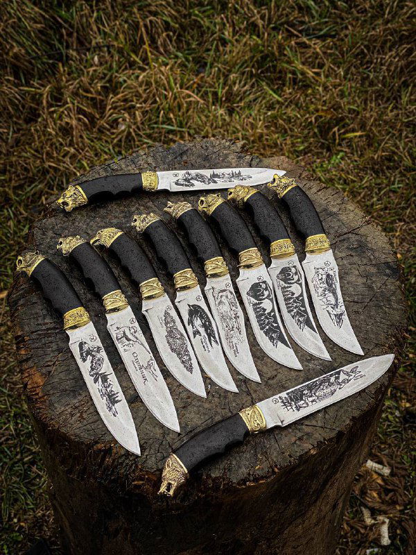 Крупный план: ряд бюджетных кизлярских ножей с гравировкой на клинках, рукояти из граба и латунными гардами, выложены на траве рядом с ножнами.