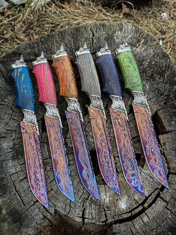Фотография ряда ножей «Пират» крупным планом: разноцветные рукояти, узорчатые дамасковые клинки и мельхиоровые гарды, фон — трава и природа