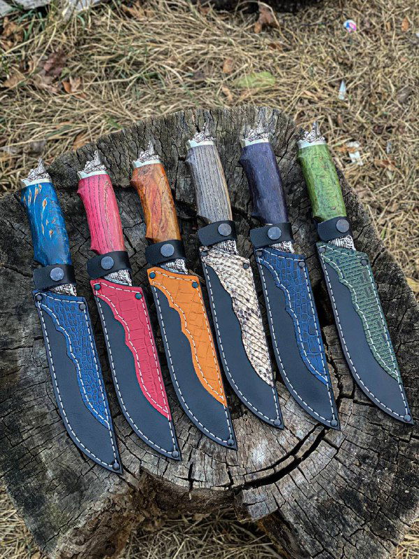Ряд ножен и рукоятей модели «Пират» с декоративной строчкой на кожаных ножнах и многоцветными рукоятями из стабилизированной берёзы, выложенные на пне