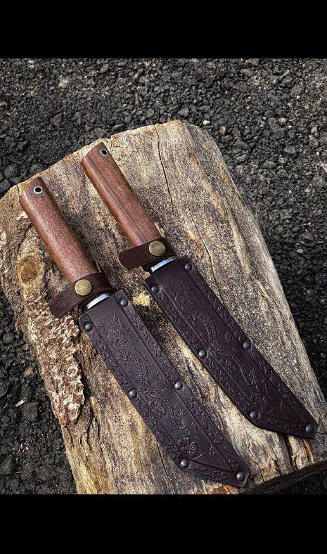 Два ножа танто в кожаных ножнах, размещённые на деревянной доске и асфальтовом фоне; показаны ножны, застёжки и оформление рукоятей из ореха.