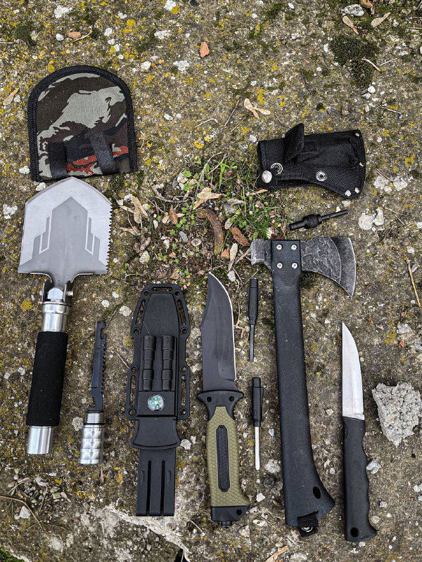 Крупный план многофункциональных инструментов: лопата, нож Gerber, топор и сопутствующие чехлы и насадки на фоне кустарника и камня.