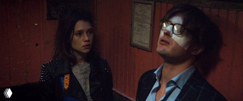 Кадр с двумя взрослыми в коридорном помещении: мужчина и женщина разговаривают у стены, тёплый свет, атмосферная сцена без привязки к лифту.