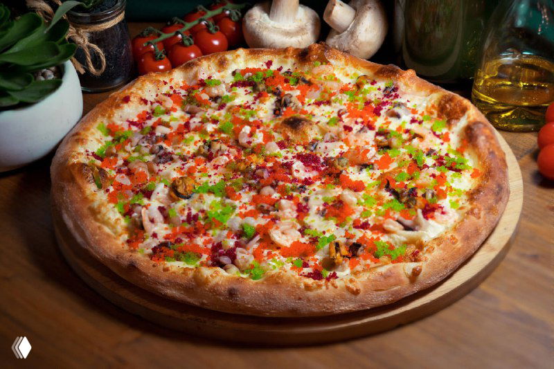 Крупная фотография пиццы на деревянной доске в кухонной обстановке: томатный соус, расплавленный сыр, кусочки колбасы, зелёный перец и приправы вокруг.