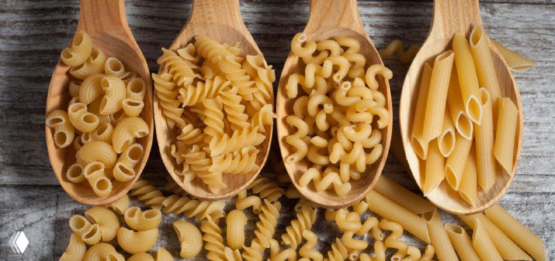 Фотография ассортимента сухих макарон: рожки, спирали, перья и пенне, уложенные на деревянных ложках на столе.