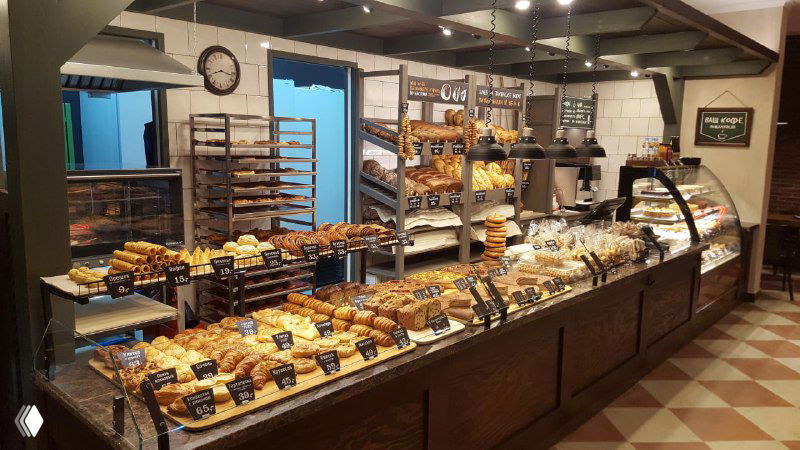 Интерьер небольшой пекарни: длинная витрина с ассортиментом хлеба, булочек и круассанов на прилавке, ценники и торговый зал на заднем плане.