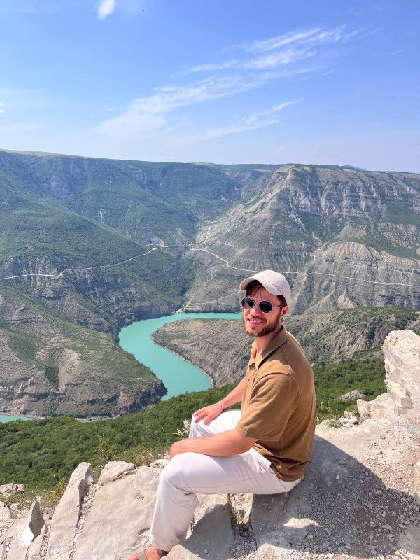 Мужчина в кепке сидит на скале над извилистой бирюзовой рекой в горном каньоне; летний пейзаж, вид с обрыва.