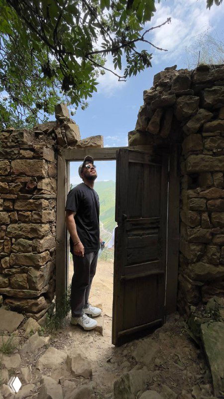 Мужчина стоит в открытой старой дверной раме среди каменных руин и смотрит вверх на небо; вокруг зелёная растительность и естественный свет.