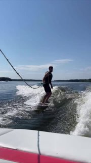 Мужчина катается на вейкборде за моторной лодкой на спокойном озере при ясном небе; видны всплески воды и силуэт райдера в движении.