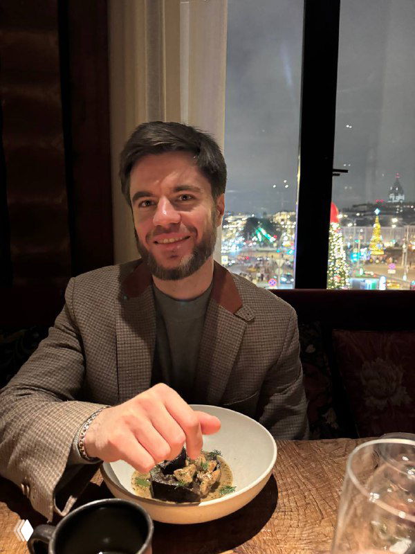 Мужчина сидит в ресторане за столом с блюдом, улыбается; на фоне видно окно с огнями ночного города и праздничной иллюминацией.