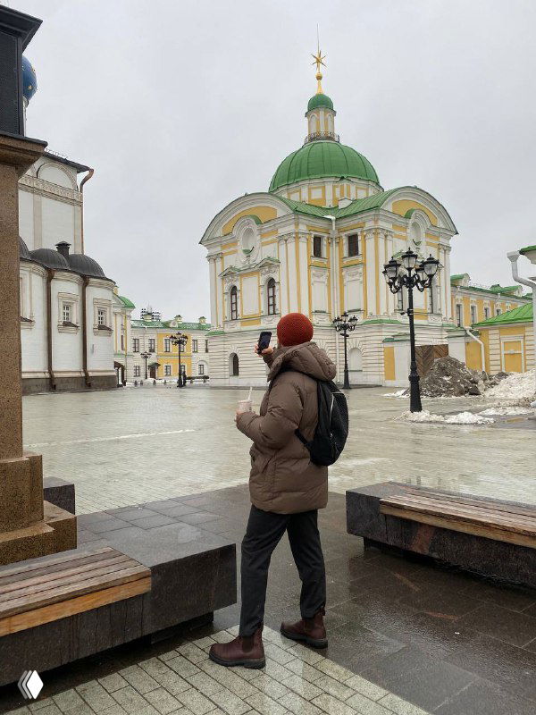 Человек в зимней куртке и рюкзаке стоит на пустой площади перед зданием с зелёным куполом; пустая мокрая улица, пасмурная погода.