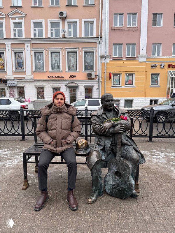 Мужчина в тёплой куртке и шарфе сидит на скамье рядом со скульптурой музыканта с контрабасом на фоне разноцветных фасадов