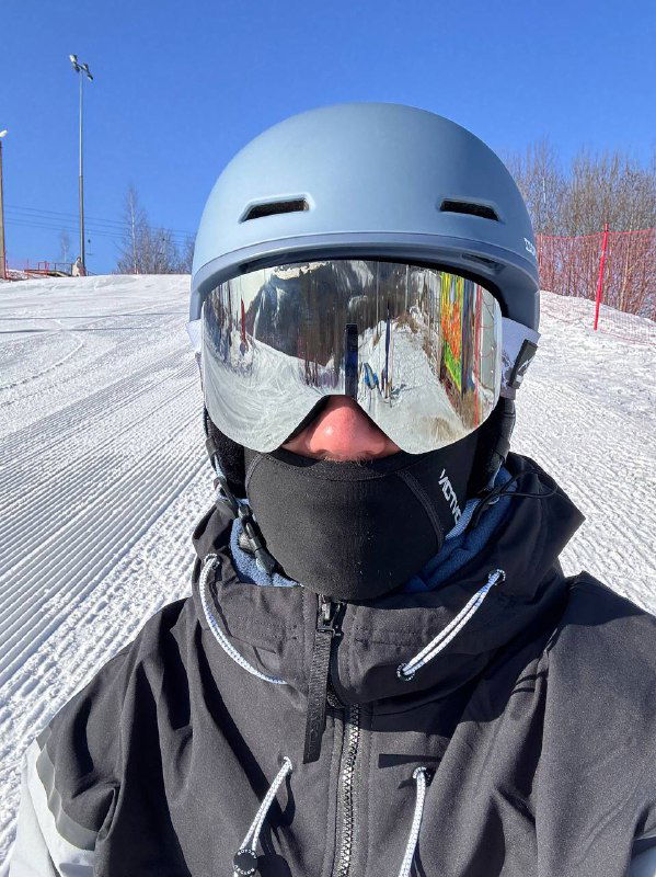 Человек в шлеме и зеркальных горнолыжных очках, лицо частично закрыто маской; на заднем плане заснеженный склон и ограждения