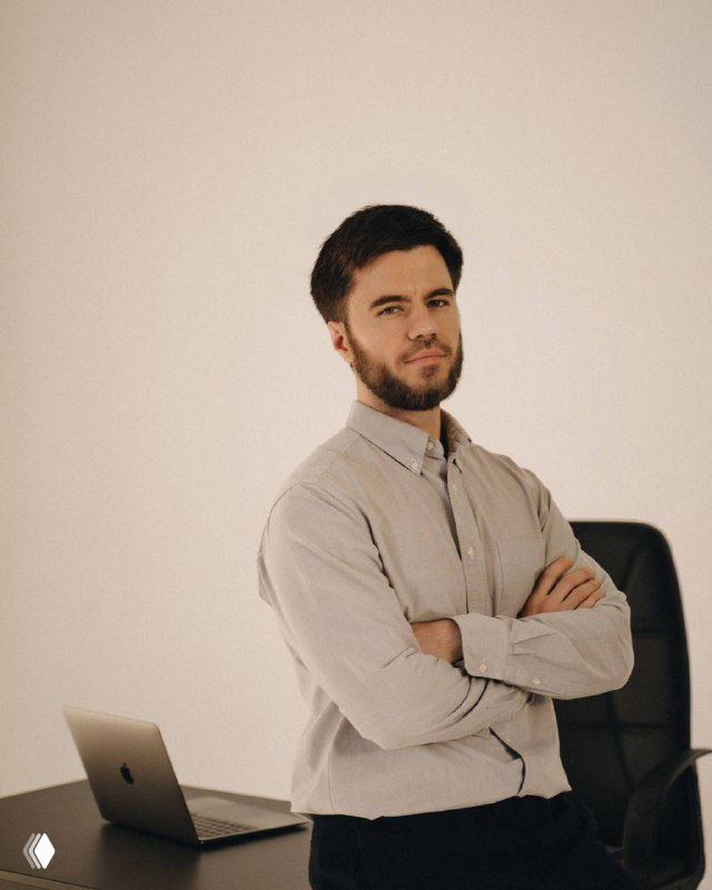 Мужской портрет: таргетолог стоит у стола с ноутбуком, руки сложены, деловой образ на нейтральном фоне, офисная атмосфера