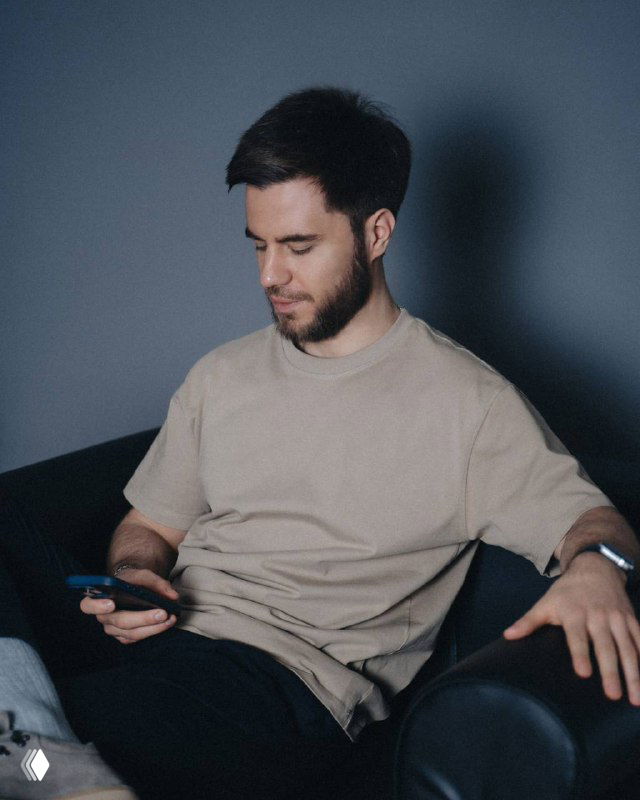 Мужчина в светлой футболке сидит в кресле и смотрит в смартфон — портрет, иллюстрирующий личность автора канала.