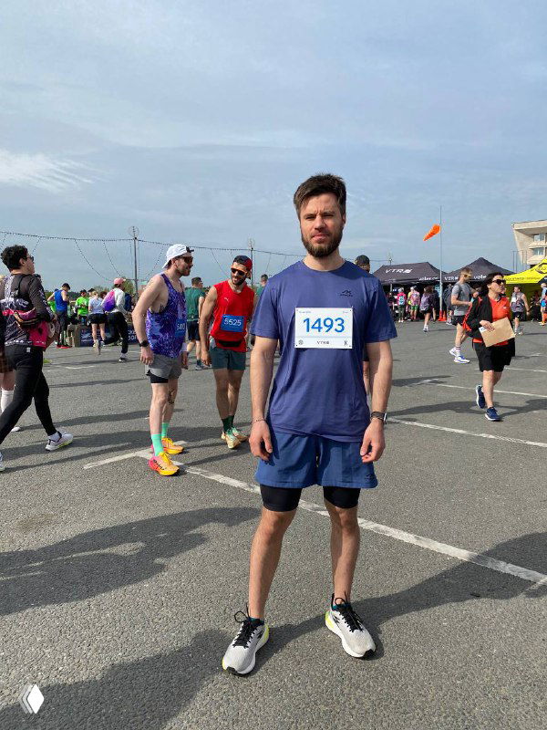 Мужчина в спортивной форме с номером 1493 стоит на асфальте перед стартом забега; вокруг другие бегуны и палатки.