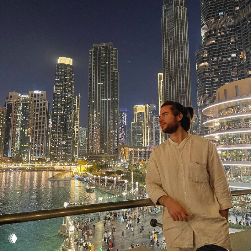 Мужчина в лёгкой рубашке опирается на перила у набережной ночью, на фоне освещённых высотных зданий, прогулочных зон и городской суеты.