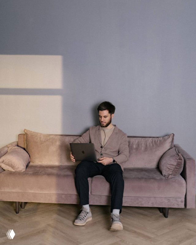 Мужчина в повседневной одежде сидит на диване с ноутбуком в светлой комнате, работает или проверяет рекламные кампании — спокойный рабочий кадр в интерьере.