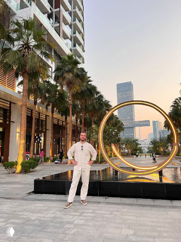 Мужчина в светлой одежде стоит у большой металлической инсталляции — кольца на набережной, на фоне пальм и высотных зданий на закате.